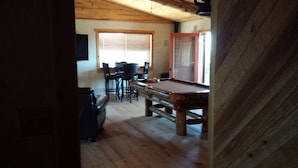 Een kamer met houten vloer, voorzien van een pooltafel, een eethoek met stoelen en een raam met jaloezieën.