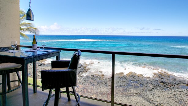 Un balcón con mesa, sillas y una lámpara colgante con vista a una playa rocosa y el océano.