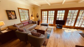 Amplia sala de estar con un sofá de cuero, dos sillones, una mesita de centro y un televisor. Amplias ventanas y puertas corredizas de vidrio permiten la entrada de luz natural y ofrecen vistas del exterior.