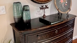 A dark wooden dresser with three drawers, a round mirror with an ornate frame, and a framed welcome sign.