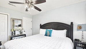 A neatly arranged bedroom with a large bed, a ceiling fan, a bedside table with a lamp, and a mirror on the wall.