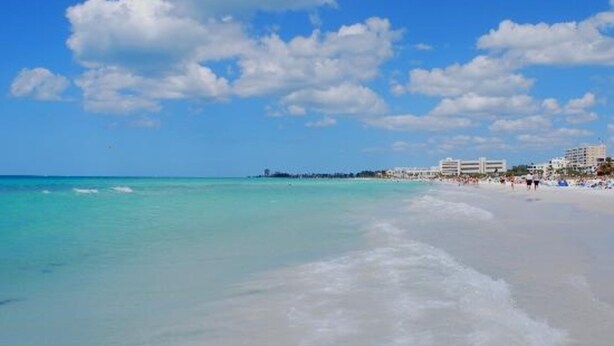 A beach with clear turquoise water, a sandy shore, and buildings in the background.