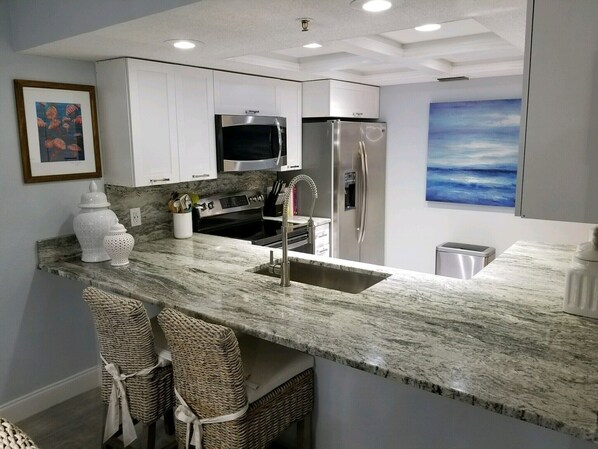 A modern kitchen with white cabinets, stainless steel appliances, a marble countertop, and wicker chairs.