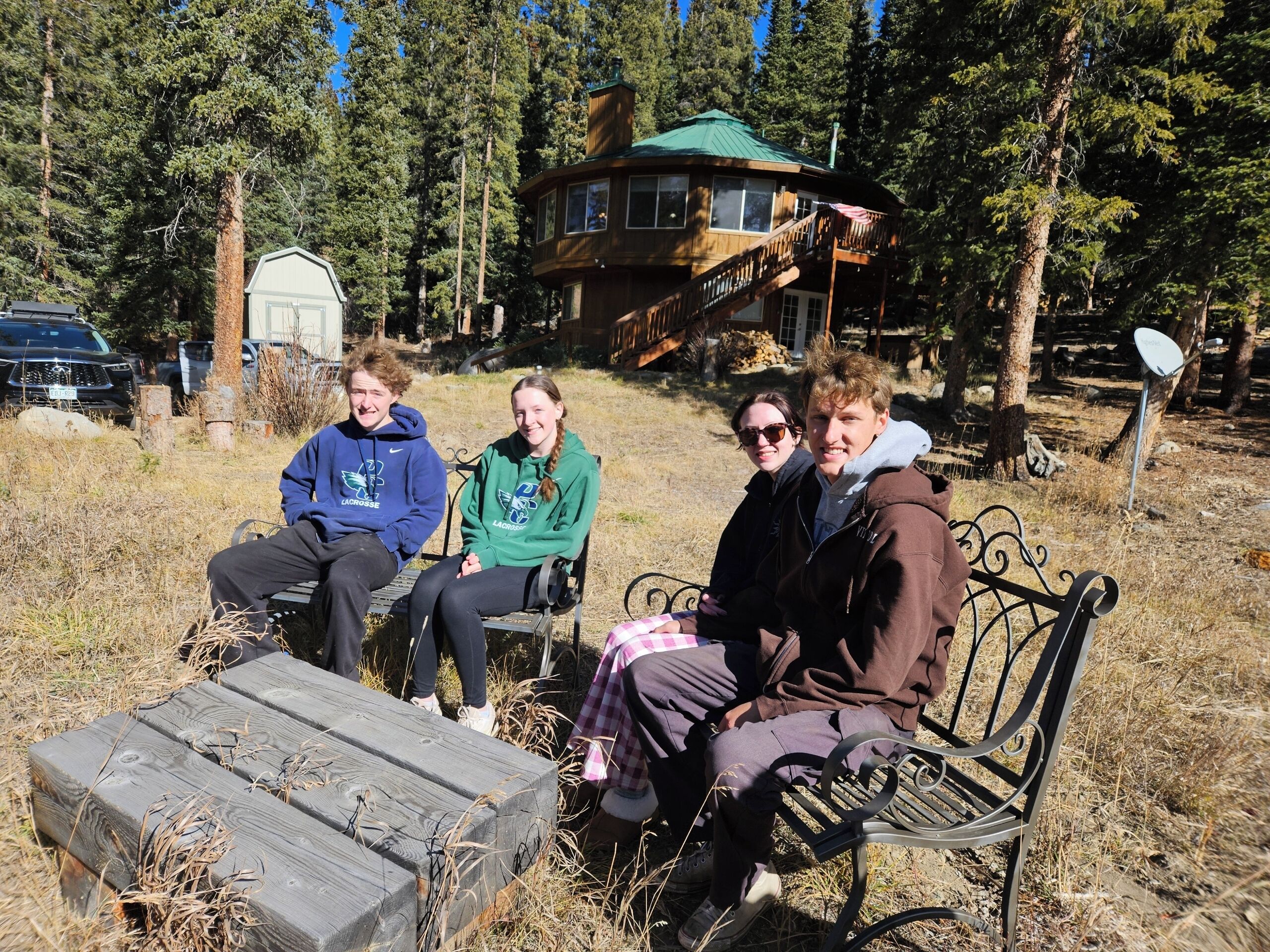 Four people sitting on a bench outdoors with a cabin in the background.