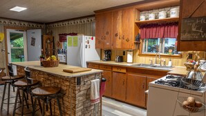 A kitchen with a stone island, wooden cabinets, and a gas stove. There are bar stools, a refrigerator, and a window with a view of the outside.