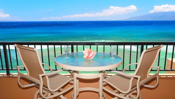 Een ronde tafel met twee stoelen op een balkon met uitzicht op een heldere blauwe zee en een berg in de verte.