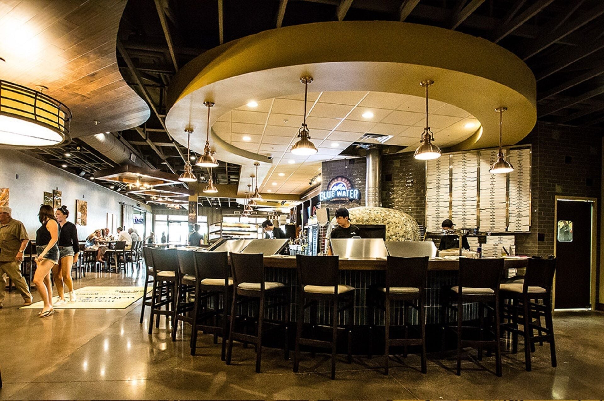 A restaurant interior with high ceilings, hanging pendant lights, and a bar area with stools.