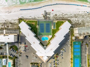 An aerial view of a property with a V-shaped building, a tennis court, a swimming pool, and a beach with umbrellas.