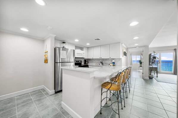 A modern kitchen with white cabinetry, a stainless steel refrigerator, and a bar area with wicker chairs. - Beach Front, Corner Unit Condominium on the 6th Floor. Incredible Beach View! (Destin)