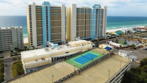 Un hôtel à plusieurs étages doté d’un court de tennis, offrant une vue sur la plage et comprenant plusieurs parkings.