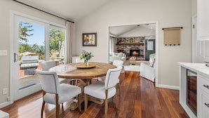 Un vaste espace de salle à manger avec une table en bois et des chaises blanches, une cuisine avec des meubles blancs et une vue sur l’extérieur.