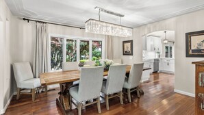 Un coin repas avec une table en bois, quatre chaises rembourrées, un lustre et une vue sur la cuisine grâce à de grandes fenêtres.