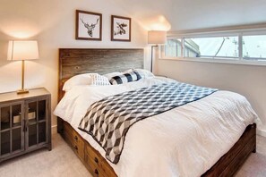 Un dormitorio con estructura de cama de madera, una mesita de noche con lámpara y ventana con vistas al exterior.