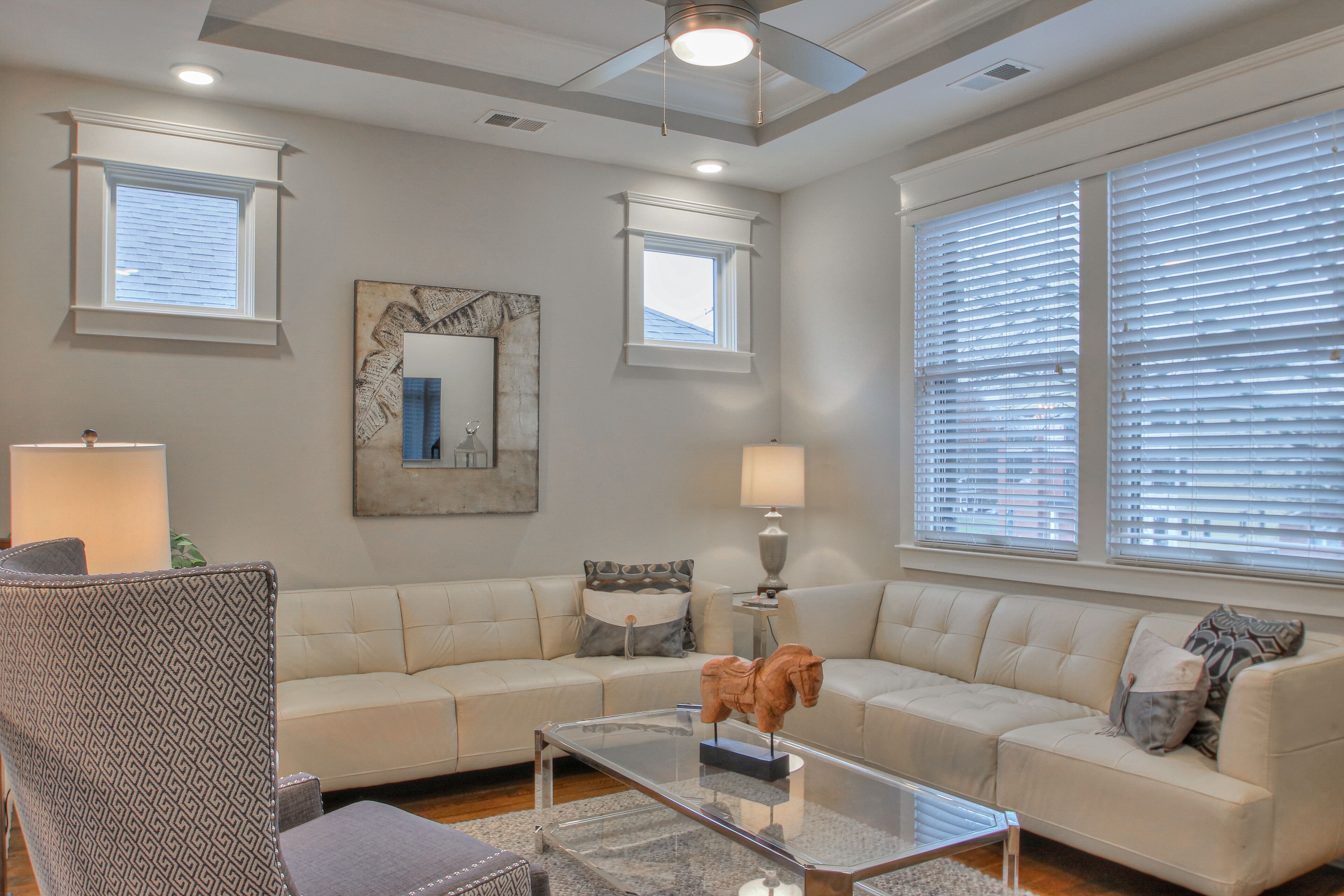 A living room with a beige sofa, a glass coffee table, and a patterned armchair. There are two windows with blinds, a ceiling fan, and a lamp on a side table.