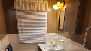 Une salle de bain avec des toilettes, un lavabo et un miroir. Il y a une fenêtre avec des stores et un porte-serviettes avec des serviettes.