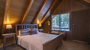 Un dormitorio con techo inclinado, vigas de madera, un ventanal con persianas, una cama con muchas almohadas, mesitas de noche con lámparas y un armario.