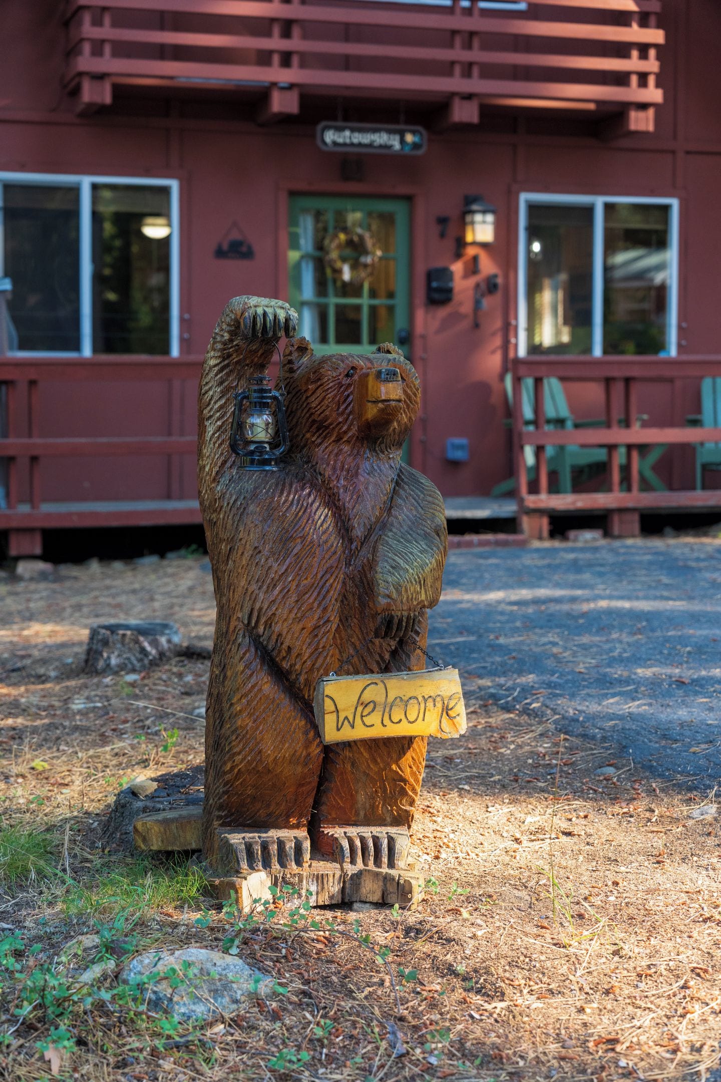 Escultura de urso de madeira segurando uma placa com a palavra 'Welcome' em frente a um edifício com fachada vermelha e porta verde.