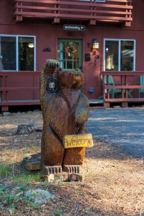 Una escultura de madera de un oso sosteniendo un cartel con la palabra 'Welcome' frente a un edificio de fachada roja y puerta verde.