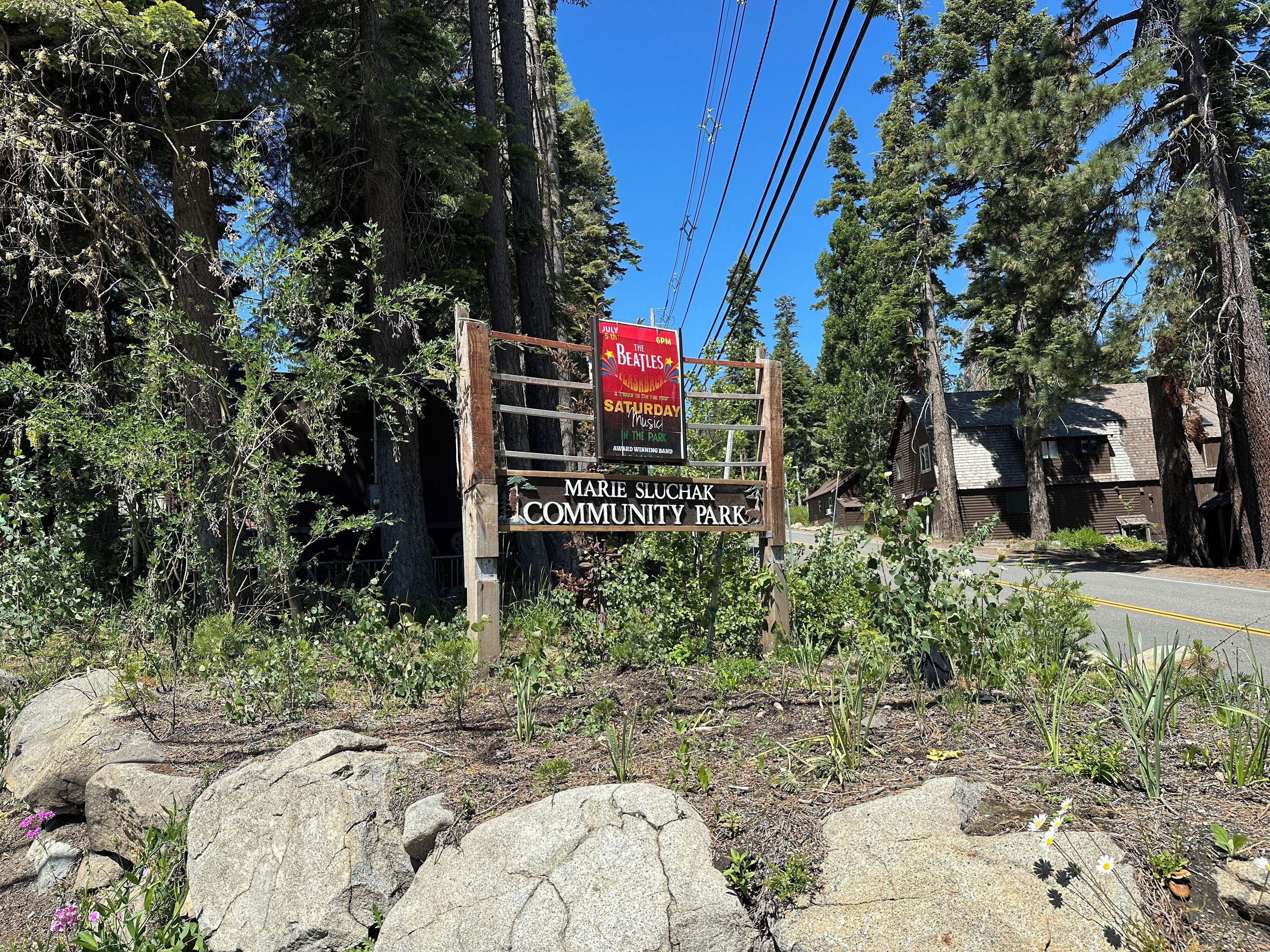 Ein hölzernes Schild mit der Aufschrift 'Marie Sluchak Community Park' und ein roter Plakatwerbung für die Veranstaltung 'The Beatles'.