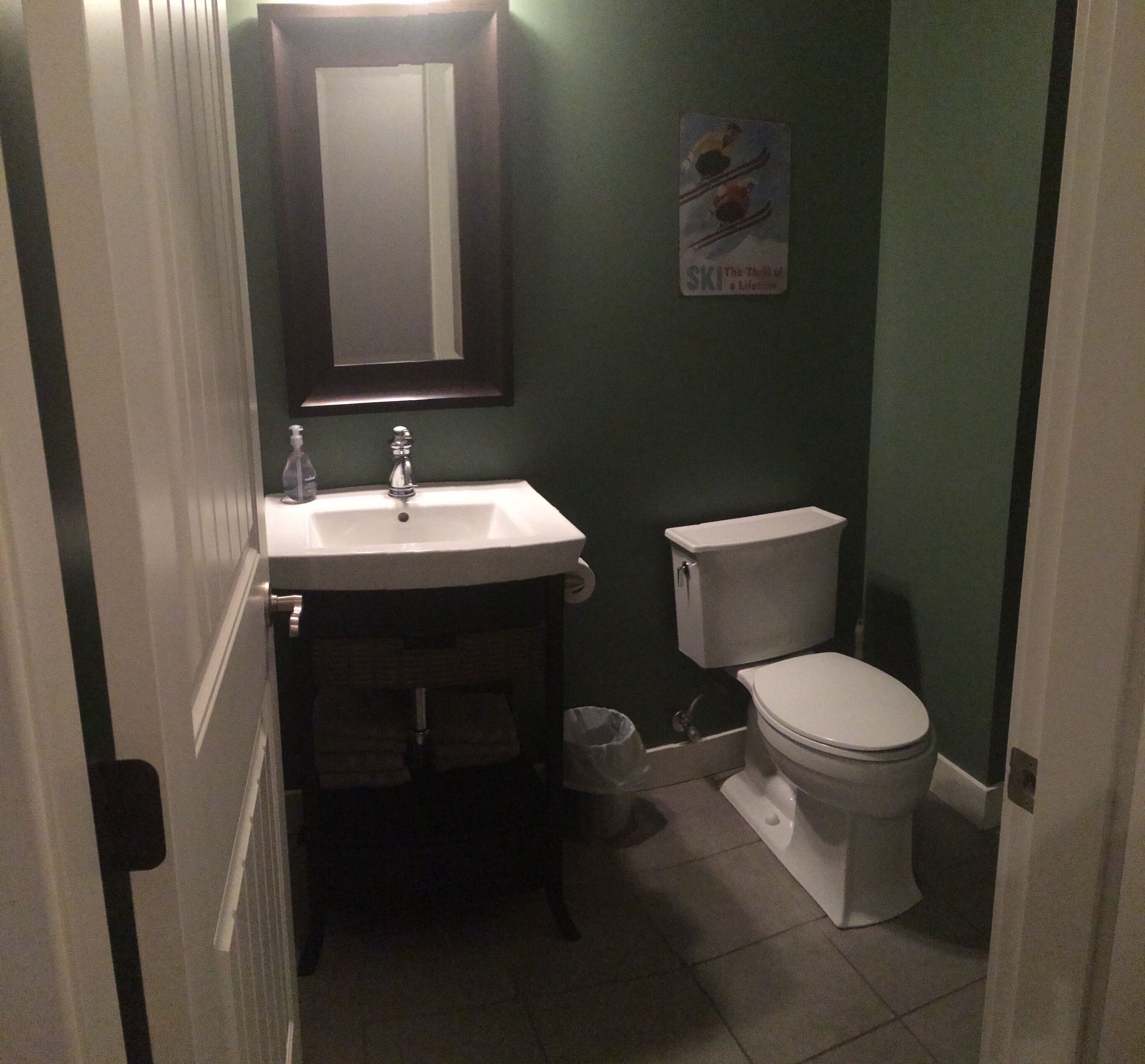 Ein kleines Badezimmer mit einem weißen Waschbecken, Spiegel und Toilette. Neben der Toilette steht ein Mülleimer.
