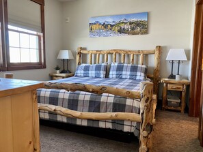 Uma cama de madeira com colcha xadrez, duas luminárias de apoio, um quadro com paisagem de montanha e uma janela com persianas.