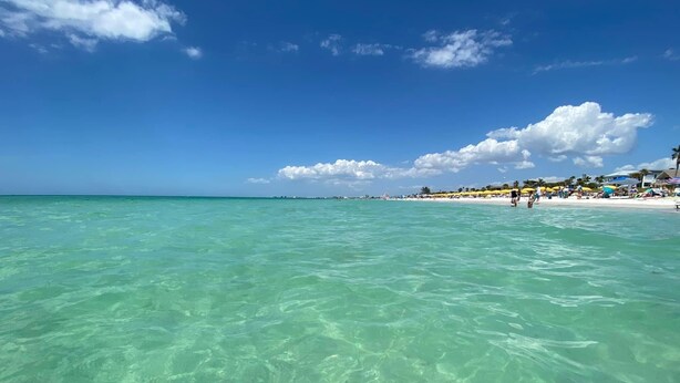A beach with clear turquoise water, a sandy shore, and a few people in the distance.