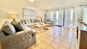A spacious living room with a sofa, armchair, and coffee table. There is a ceiling fan, a lamp, and a large window with curtains.