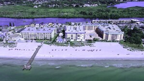 An aerial view of a coastal hotel with multiple buildings, a pier extending into the water, and a beach with umbrellas.
