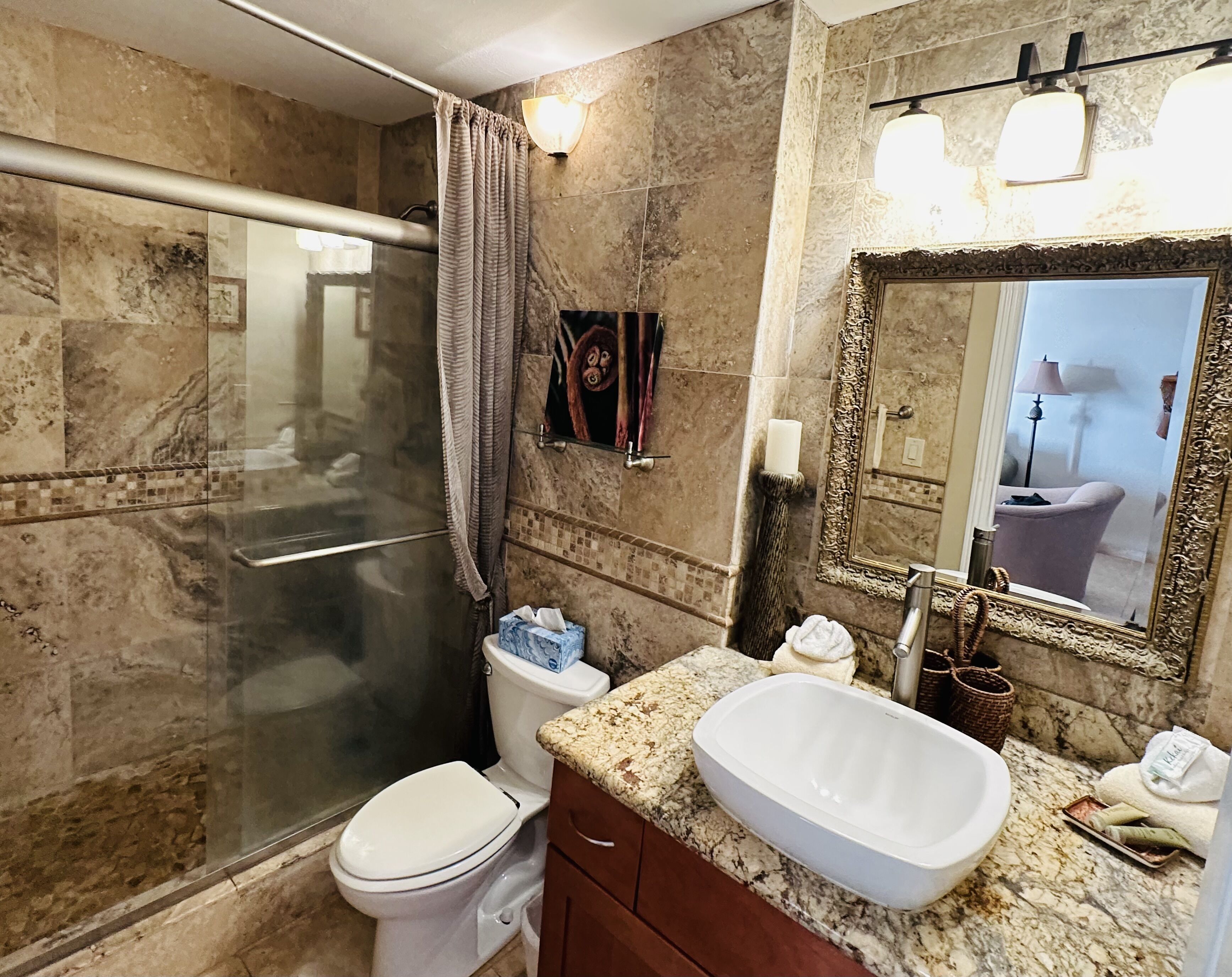 Un baño con ducha envuelta en vidrio, inodoro blanco, lavamanos de mármol y un espejo grande con marco decorativo.
