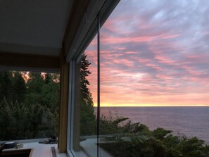 Una vista al atardecer desde un edificio moderno con amplios ventanales que dan a un bosque y al océano.