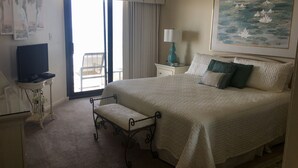 Habitación de hotel con una cama grande, un televisor sobre una mesita auxiliar, una silla junto a la ventana y un cuadro en la pared.