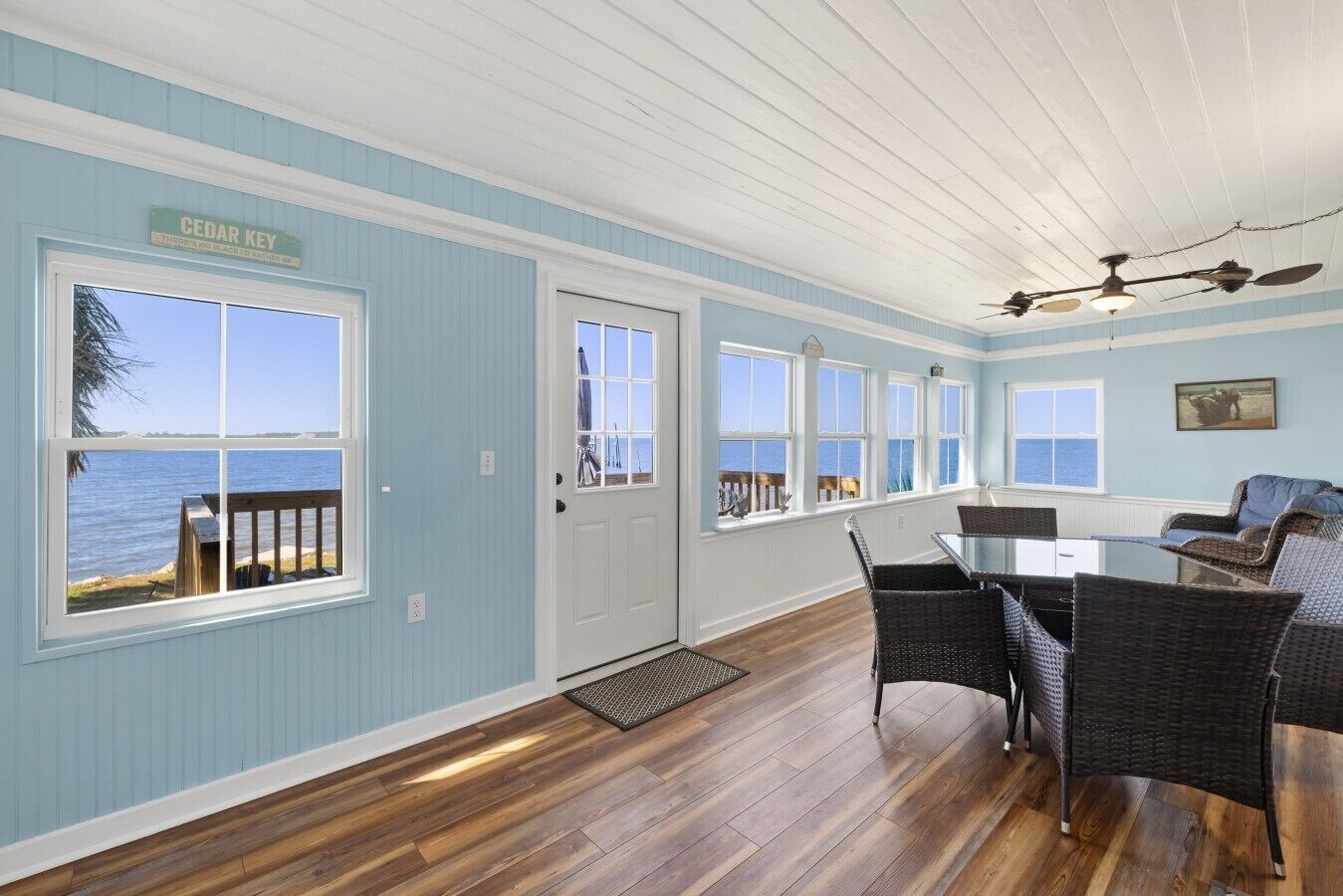 A room with a view of the water, a door with a sign, wicker furniture, and a ceiling fan.