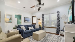 Un salon avec un canapé bleu, des fauteuils beige, un pouf blanc, un ventilateur de plafond, un téléviseur et une fenêtre avec des stores.