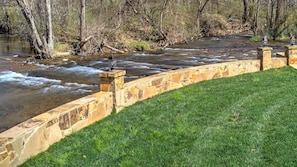 Eine steinerne Stützmauer neben einer grünen Fläche an einem Fluss, umgeben von Bäumen.