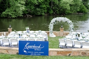 Hochzeitsdekoration mit weißen Stühlen und Tischen, einem blauen Schild und einem Blick auf einen Fluss und Grünflächen.