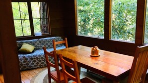 A wooden dining table with four chairs, a patterned sofa with cushions, and a window with a view of greenery. - The Treehouse - a Romantic Russian River Retreat (Guerneville)