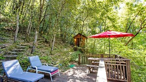 A wooden deck with lounge chairs and a red umbrella, surrounded by a forested area with a cabin.