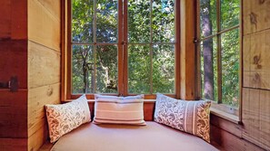 Une chambre dans un chalet en bois, dotée d’une grande fenêtre, d’un lit agrémenté d’oreillers à motifs et offrant une vue sur une forêt.