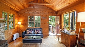 L’intérieur d’un chalet accueillant, avec un plafond en bois, un canapé avec des coussins, une lampe, un mur en pierre et une fenêtre donnant sur la verdure.