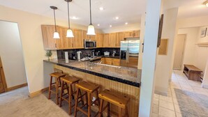 Una cocina con armarios de madera, encimera de granito, microondas, estufa y refrigerador. Hay tres taburetes de bar de madera y lámparas colgantes sobre la encimera.