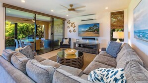 A modern living room with a sofa, coffee table, and TV. There is a sliding glass door leading to a balcony with outdoor furniture. - Designer - Best View!Ground Flr 2 Bdm/2Bth 39c! Remaining Fall$400 (Kihei)