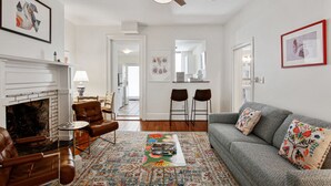 A living room with a fireplace, leather armchair, grey sofa, and a colorful rug. There are two bar stools and a ceiling fan. - Colonial Park Retreat! Historic District Prime Location! (Savannah)