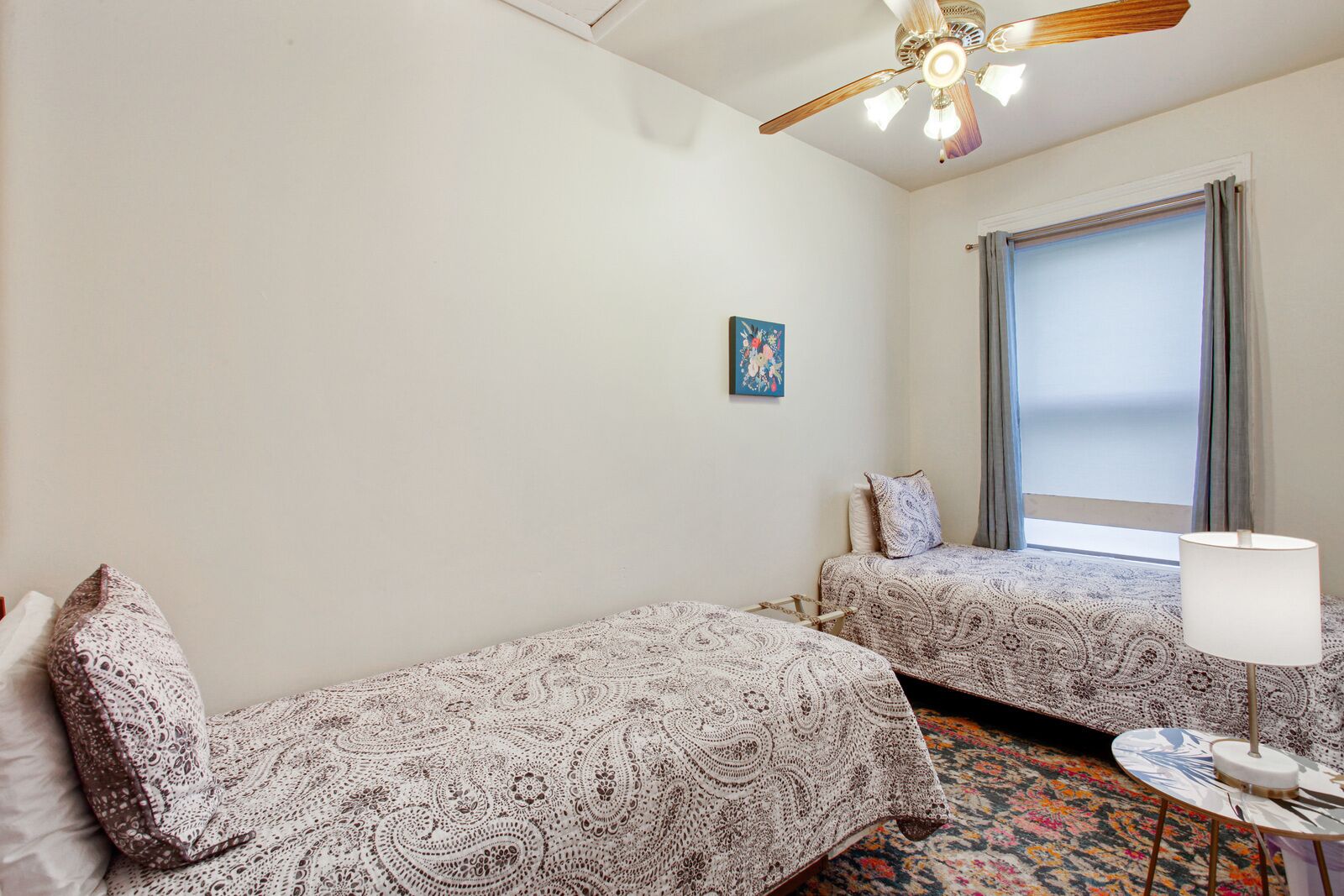 Une chambre avec deux lits, un ventilateur de plafond, une fenêtre avec des rideaux et une petite table avec une lampe.