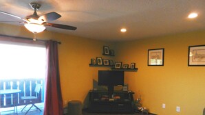 Un salon doté d’un ventilateur de plafond, d’un téléviseur à écran plat, d’une table basse et d’un canapé. Des tableaux encadrés ornent le mur, et une fenêtre est agrémentée de rideaux.