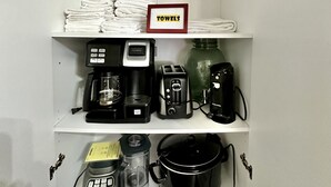 Un estante blanco con toallas blancas dobladas ordenadamente, un letrero que dice “TOWELS” y diversos electrodomésticos de cocina, como una cafetera, un tostador y una olla de cocción lenta.