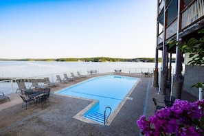 A swimming pool with lounge chairs and tables, surrounded by a white fence, with a view of a lake and distant land. - A COZY GETAWAY!Spectacular Main Channel Wtrfrt Condo Walkin Level 2BR-Unit 10210 (Osage Beach)