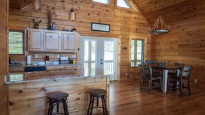 A wooden cabin interior with a kitchen area featuring cabinets, a dining table with chairs, and a bar stool. - Brooke's Place Deep Creek Area in Bryson City- Luxury Cabin (Bryson City)