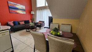 Een moderne woonkamer met een bank, fauteuils, een eettafel met stoelen, een televisie en een schilderij aan de muur.