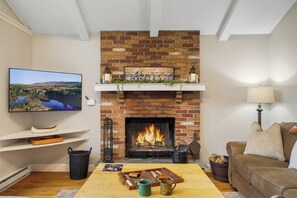 A living room with a brick fireplace, a television, a couch, and a coffee table. - Sleeps 14! Wooded Retreat, Prime Location, Lake Access With Hot Tub (Lake Harmony)