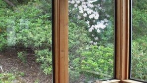 A room with a wooden table and a large window overlooking a garden with various plants and flowers. - Frank's Place: Classic Inverness  Home with Bay Views, Hot Tub (Inverness)